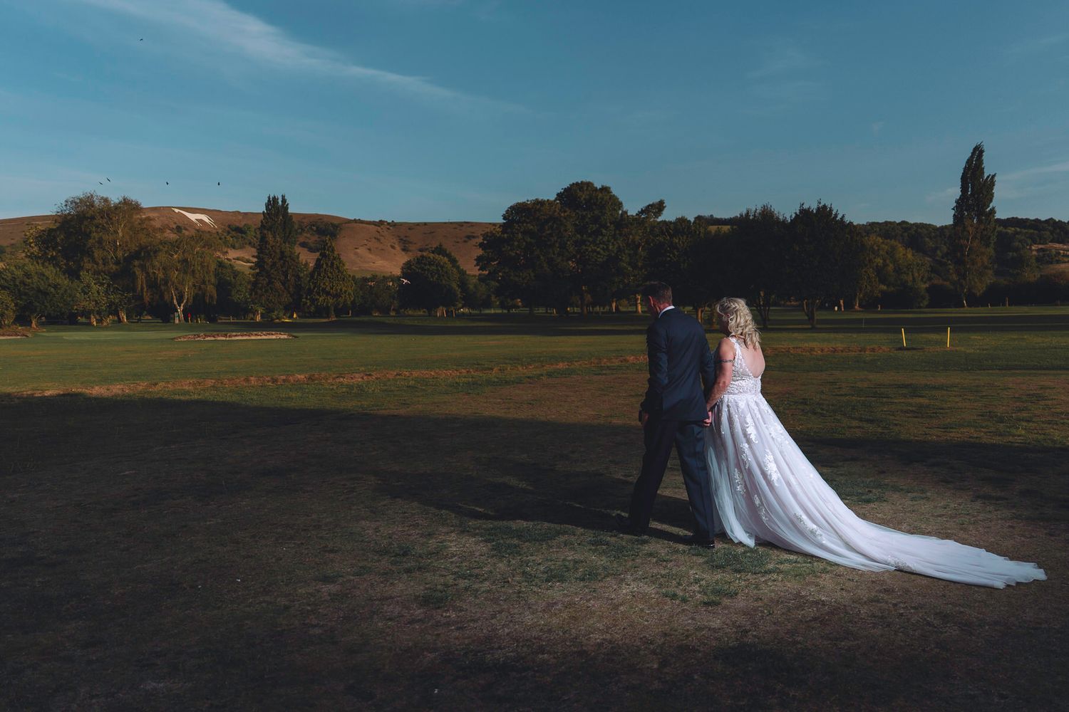 A bride and groom walk hand in hand across a sunlit green field with rolling hills and trees in the background.