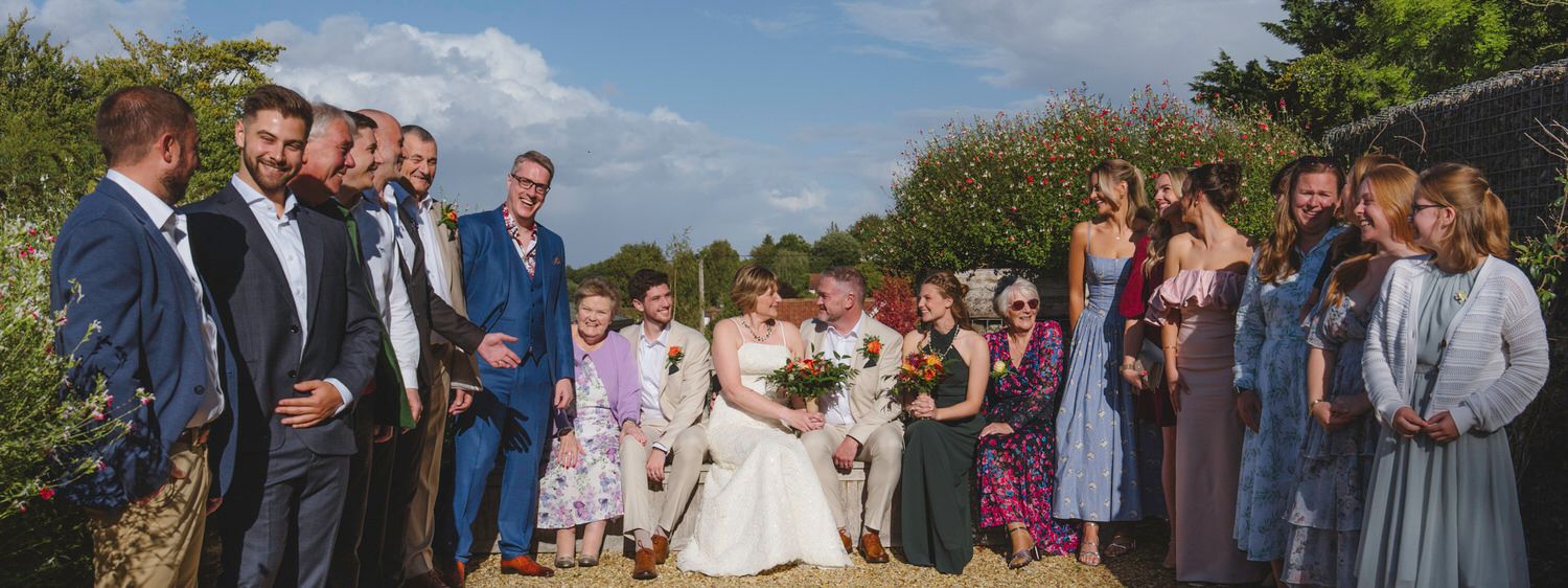 A bride and groom pose with their wedding party outdoors, surrounded by guests in formal attire on a sunny day.