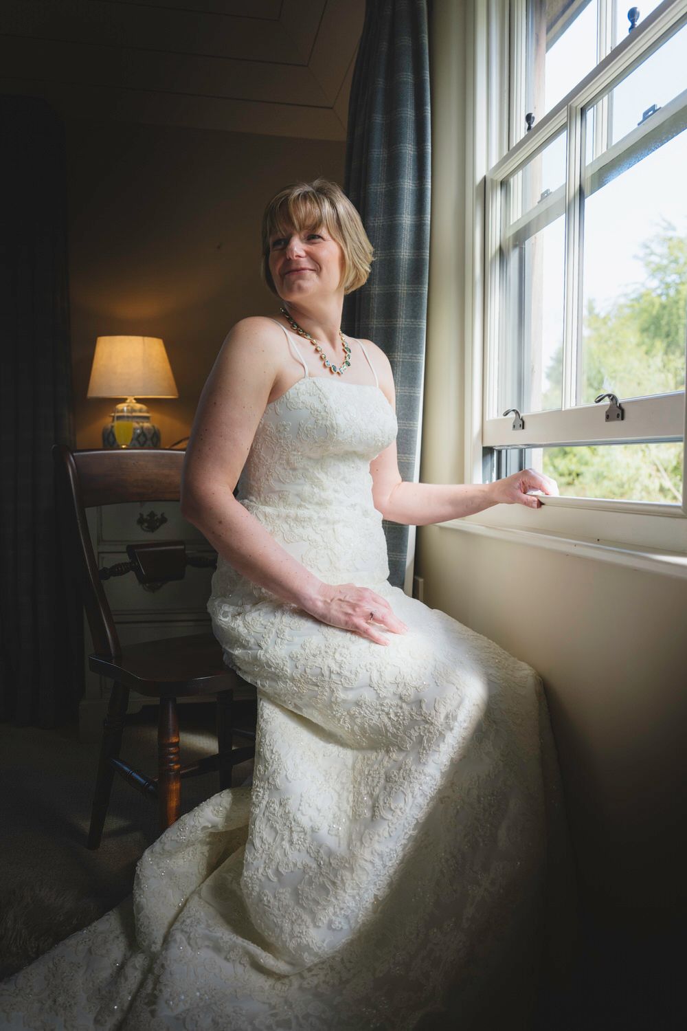 Bride in white lace strapless gown sitting by window with natural light, wearing pearl necklace in elegant room.