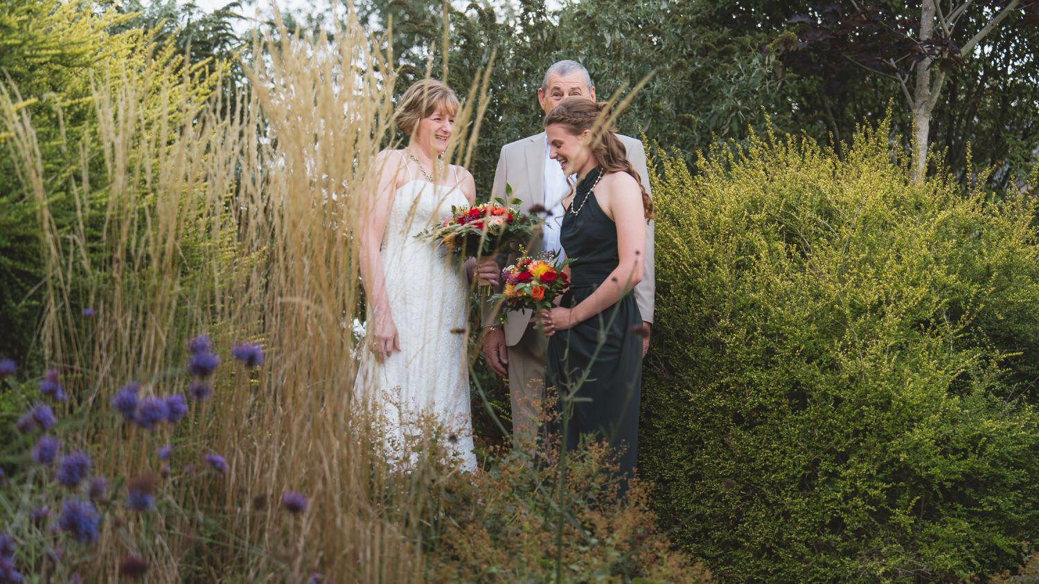 Bride and groom stand with officiant among tall grasses and wildflowers in a lush garden wedding ceremony.