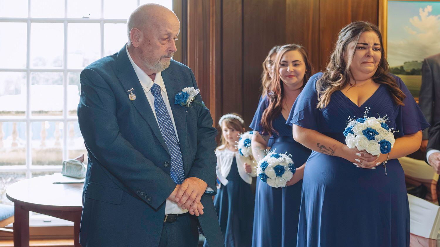 Wedding party with bridesmaids in blue dresses holding bouquets standing beside an elderly gentleman in a suit.