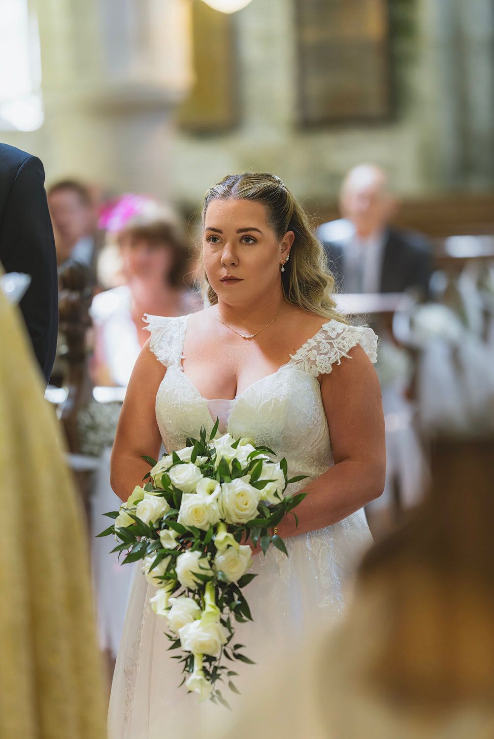 Bride in lace wedding dress holding white rose bouquet walking down church aisle.