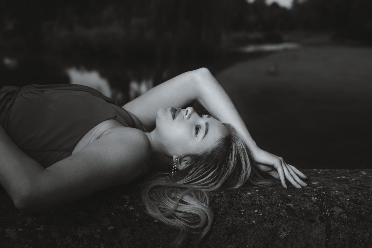 Black and white photo of a young blonde woman lying on a rock by a lake, head tilted back dreamily.