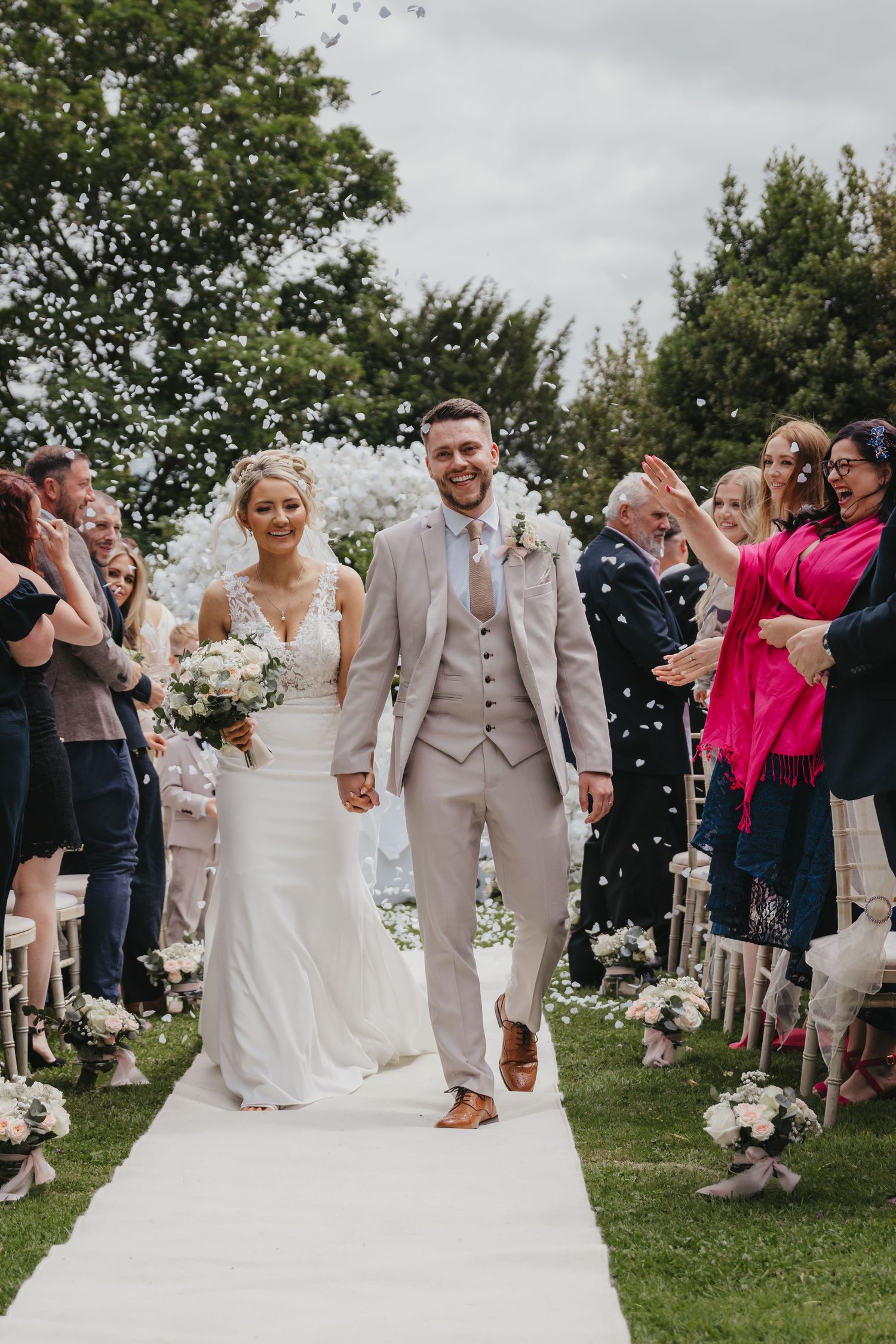 Bride and groom walk down outdoor aisle as guests throw white confetti at a garden wedding ceremony.