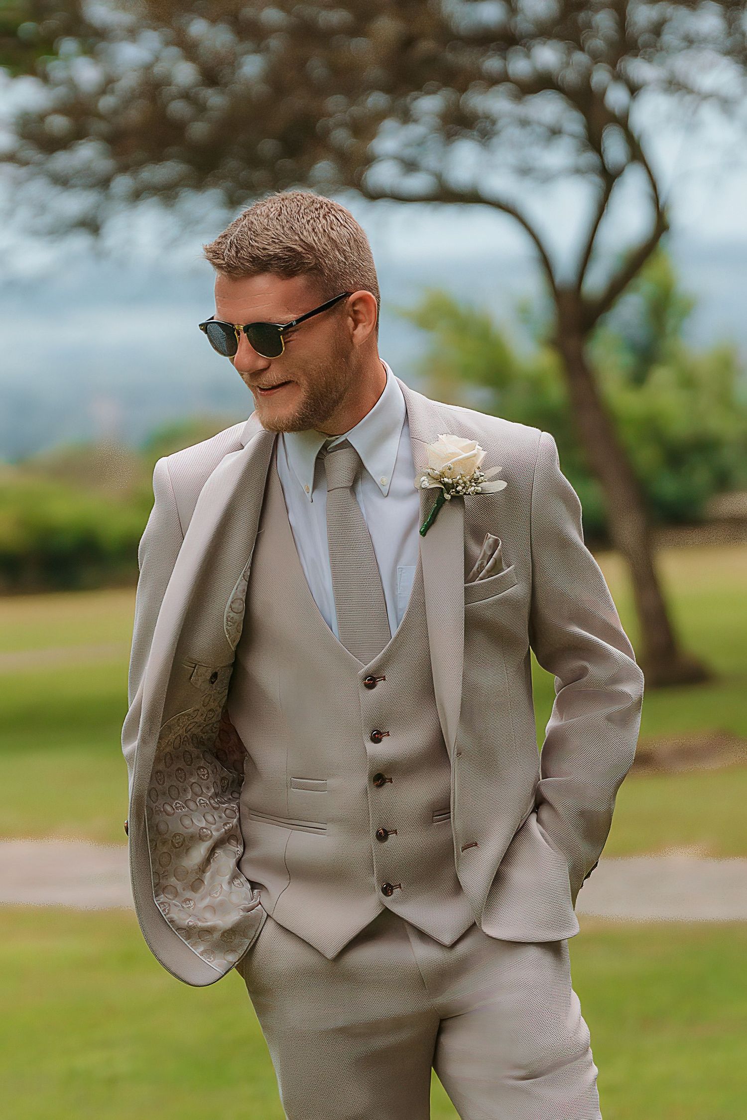 Groom in beige three-piece suit with boutonniere and sunglasses standing outdoors on a sunny wedding day.