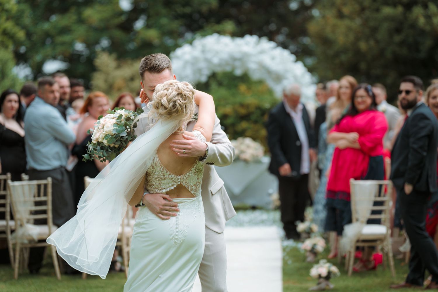 Bride and groom share a romantic embrace during outdoor wedding ceremony with floral arch and guests in background.