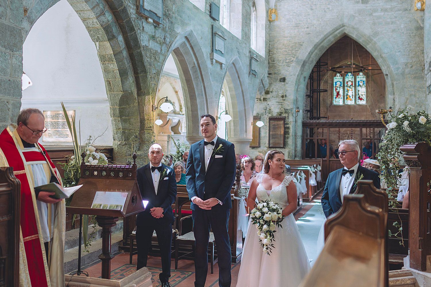 Bride and groom stand at the altar during a church wedding ceremony, surrounded by guests and floral decorations.