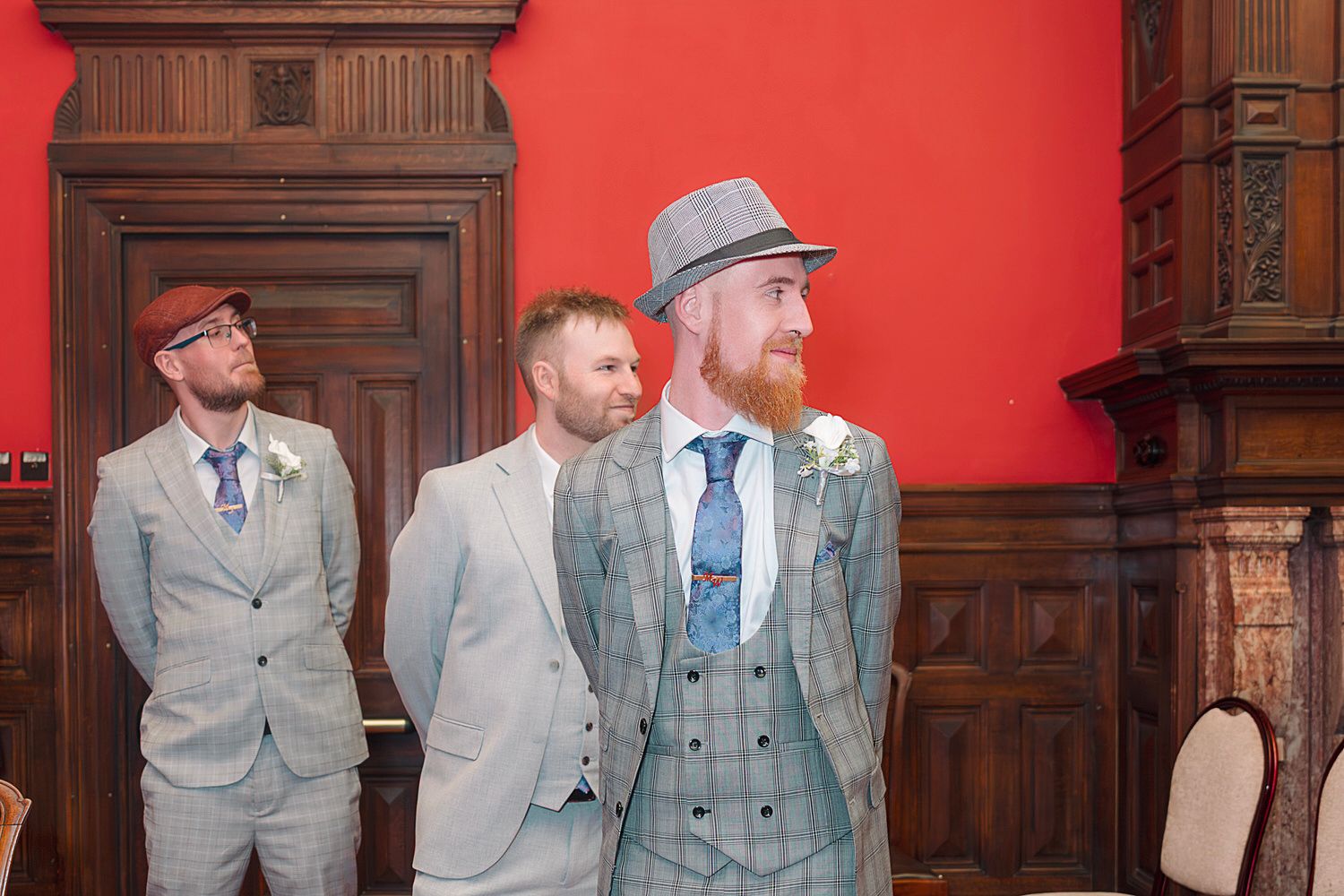 Three groomsmen in grey checked suits and hats stand in a red-walled wedding venue.