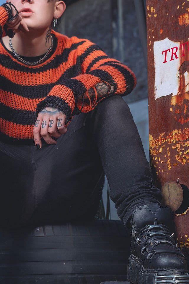 Person in orange and black striped knit sweater with hand tattoos, black jeans and boots, seated outdoors.