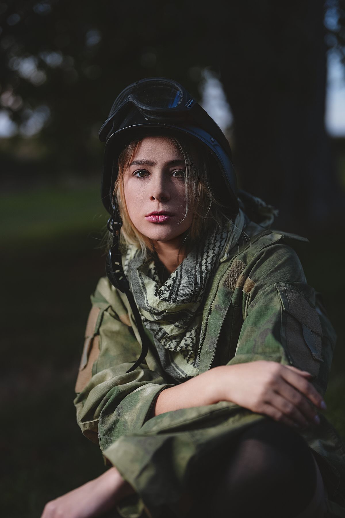 Woman in camouflage military jacket and black helmet with goggles sitting outdoors at dusk.