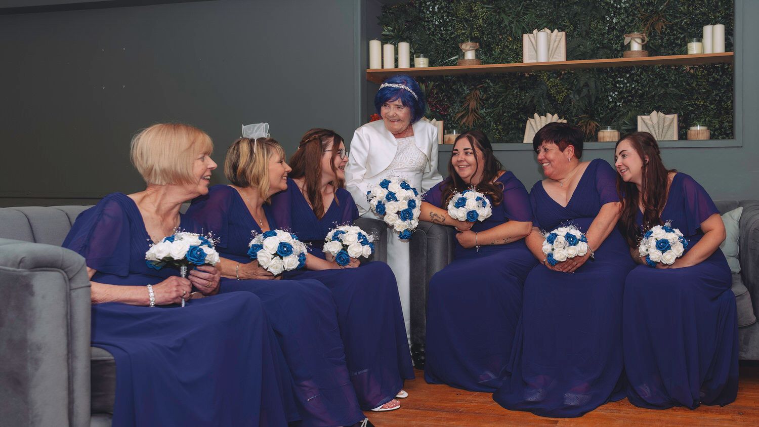 Bride in white gown with bridesmaids in royal blue dresses holding teal and white bouquets at a wedding venue.