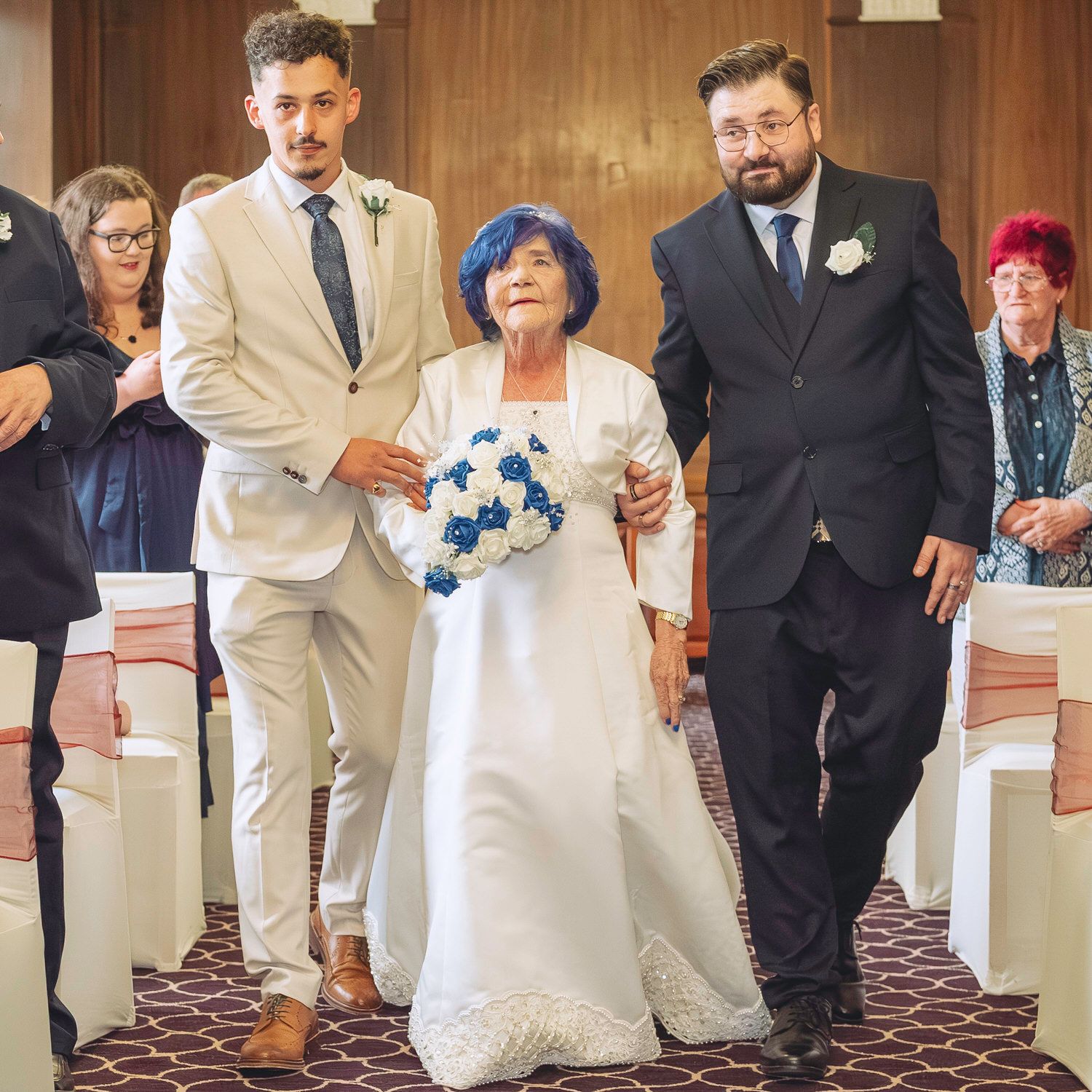 Elderly bride in white gown walks down the aisle escorted by two men in suits at a wedding ceremony.