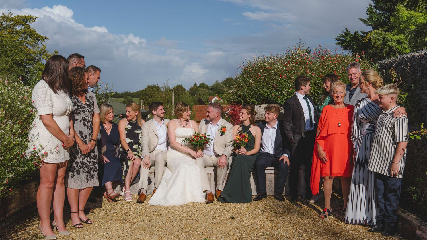 Wedding party poses outdoors on a sunny day, bride in white gown surrounded by guests in colorful attire.