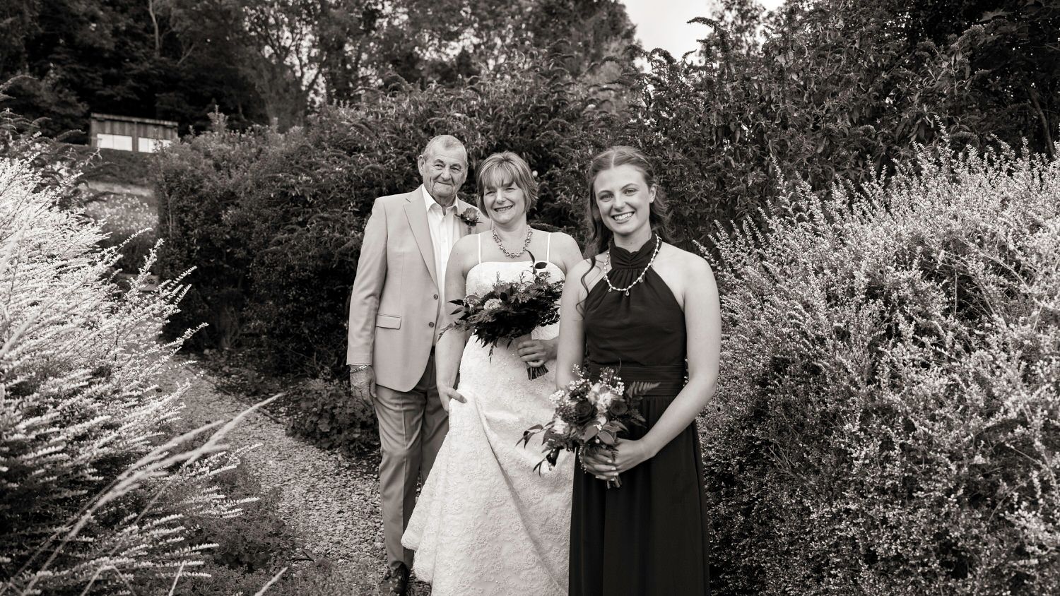 Black and white wedding photo of bride, groom, and bridesmaid posing outdoors among lush greenery.