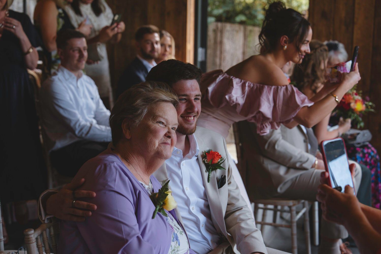 Groom smiling with mother at wedding reception, surrounded by guests in a warmly lit venue.