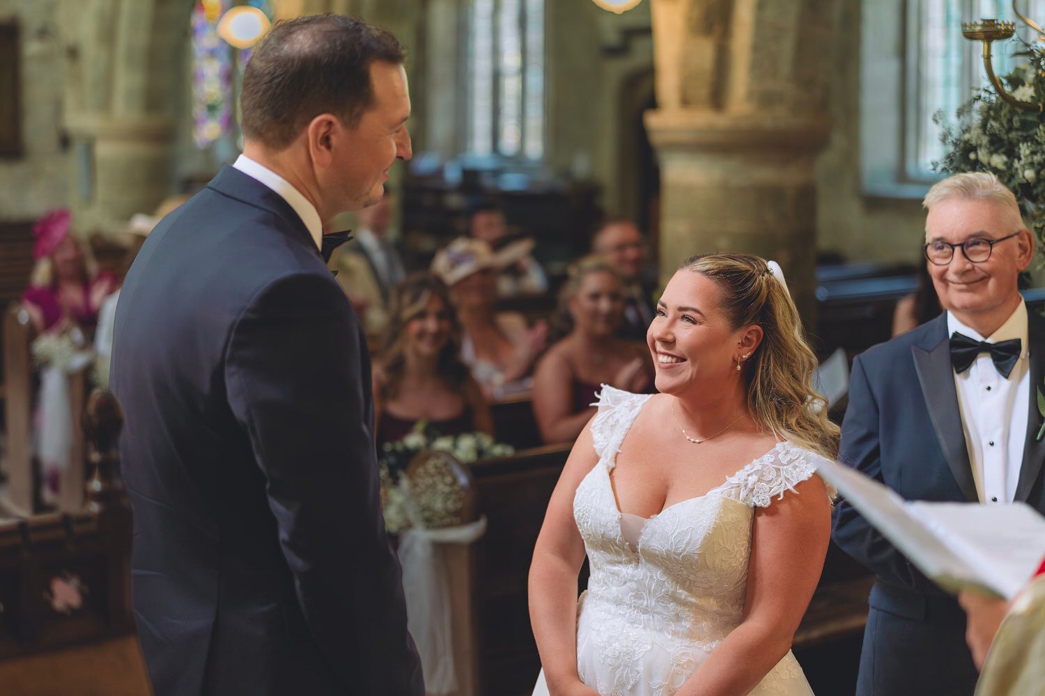 Bride smiling joyfully at groom during wedding ceremony in a historic church with guests in background.