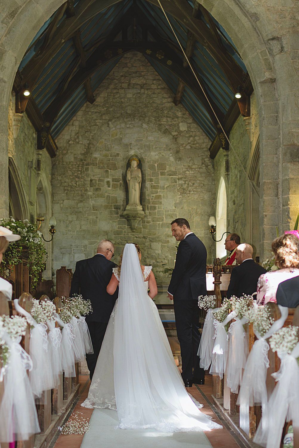 Bride and groom exchange vows at stone church altar, pews decorated with white tulle and baby's breath flowers.