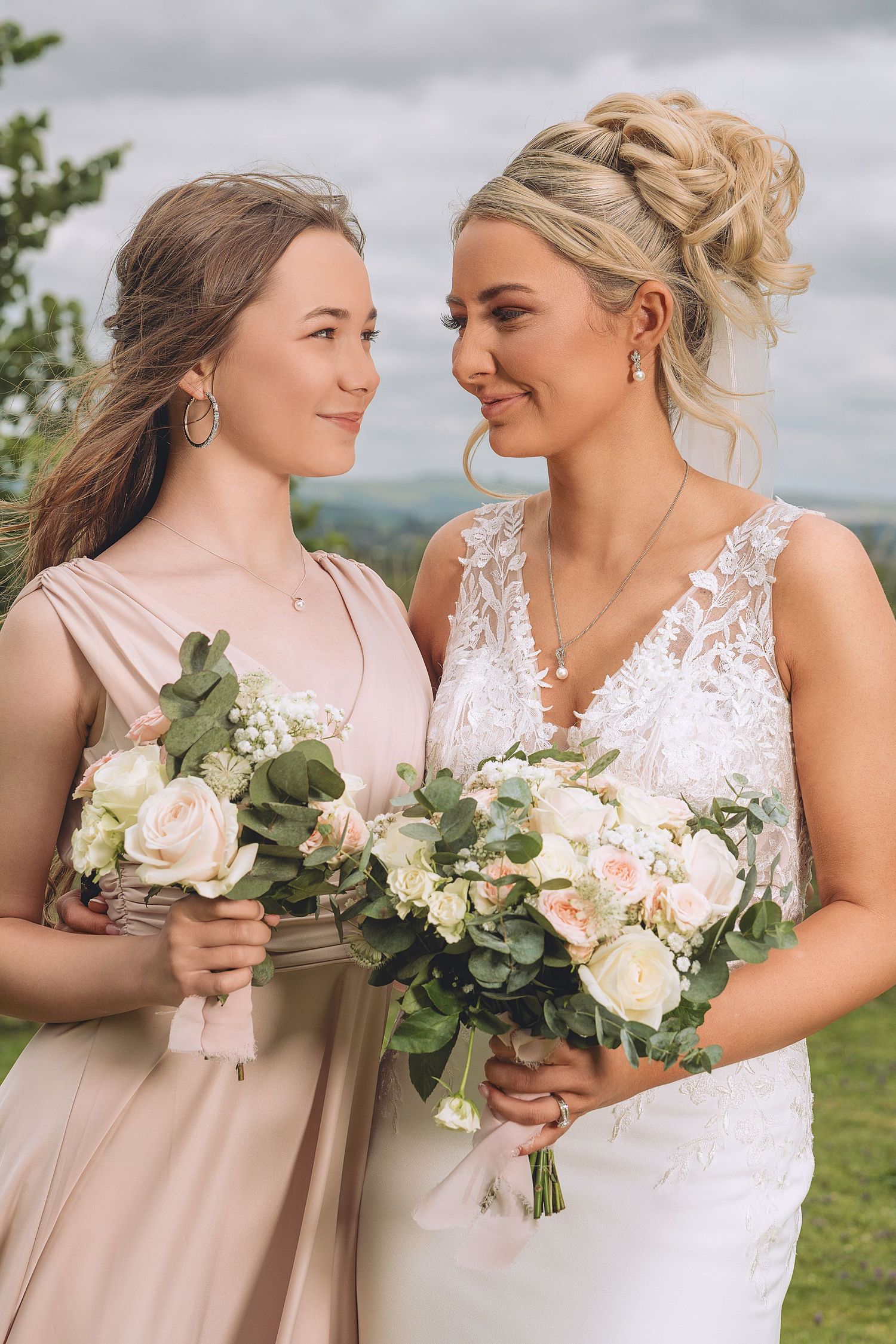 Bride in white lace gown and bridesmaid in blush dress holding bouquets of roses and eucalyptus outdoors.