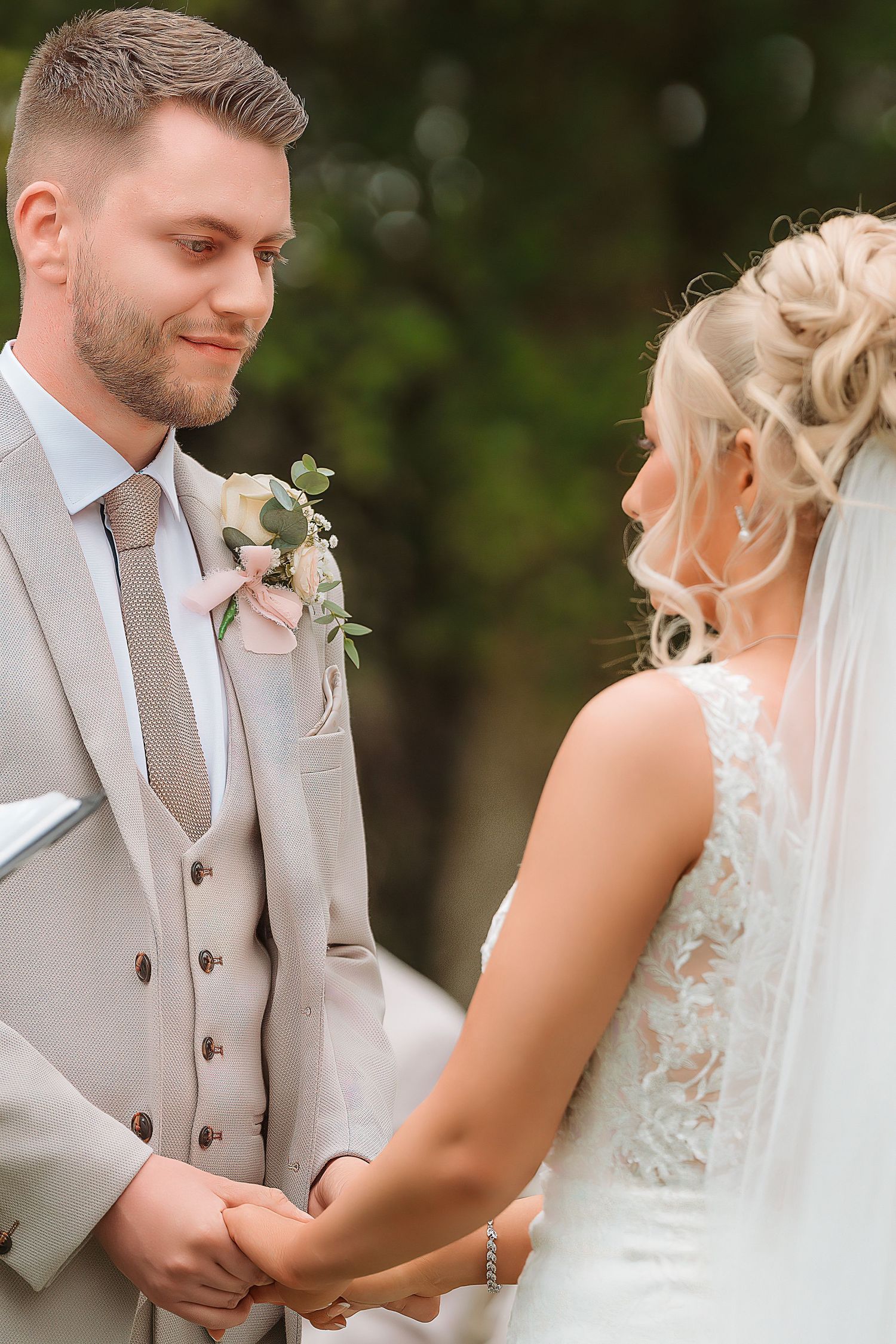 Groom in beige suit holds brides hands at outdoor wedding ceremony, exchanging vows with tender smiles.