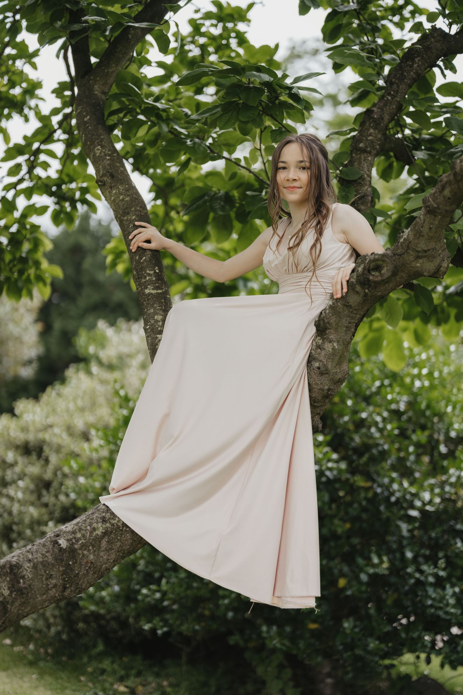 Young woman in a blush pink maxi dress posing gracefully in a tree surrounded by lush green foliage.