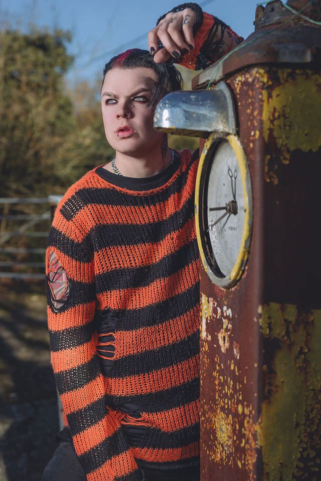 Young man in orange and black striped distressed sweater leaning against a vintage rusty gas pump outdoors.