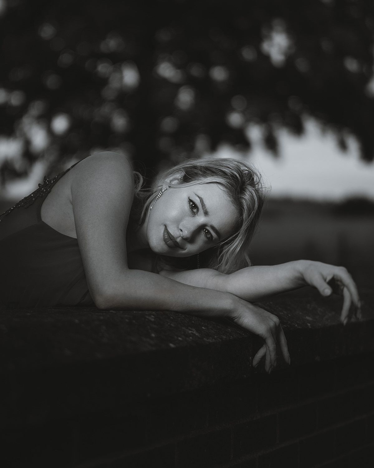 Black and white portrait of a young blonde woman leaning on a surface outdoors with bokeh tree background.