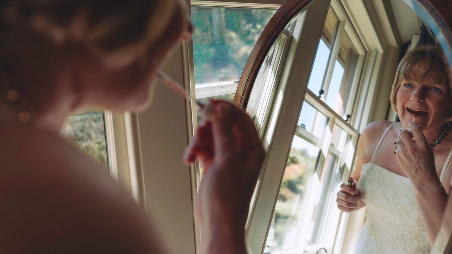 Bride applying makeup while smiling at her reflection in an oval mirror on her wedding day.