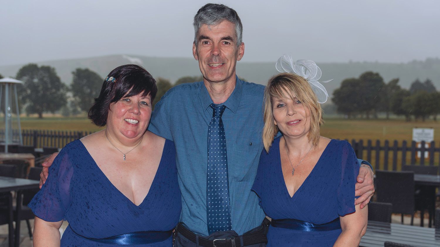 Three people in blue formal attire smiling together outdoors at an event with green hills in the background.