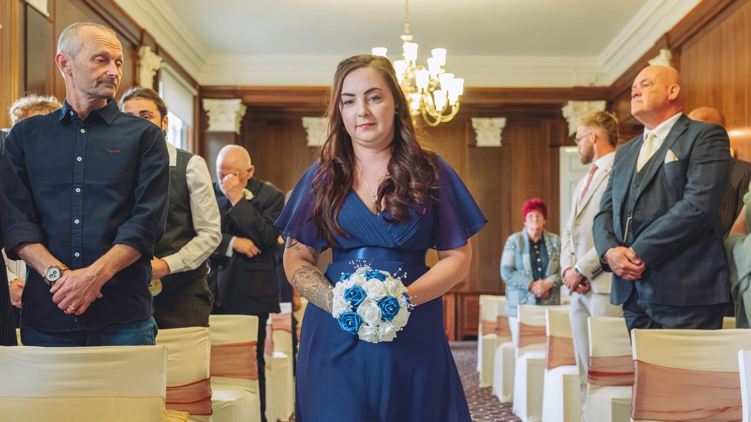 Woman in blue dress holding bouquet walks down wedding aisle in elegant ceremony hall with guests standing.