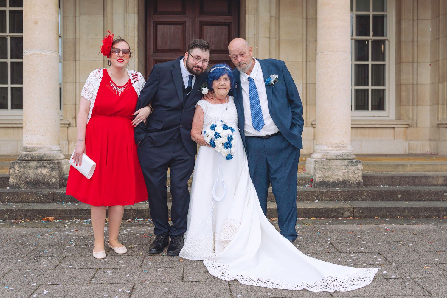Wedding party of four pose outside venue, bride in white gown, groom and guests in navy and red attire.