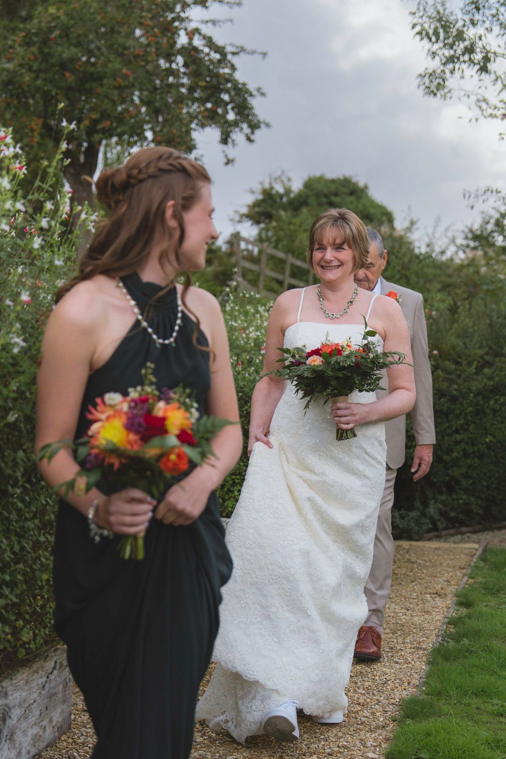 Bride in white lace gown holding bouquet walks garden path with groom, greeted by bridesmaid in black dress.