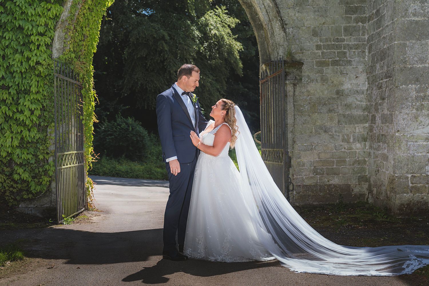 Romantic couple share intimate moment under ivy-covered stone archway with cathedral veil flowing.