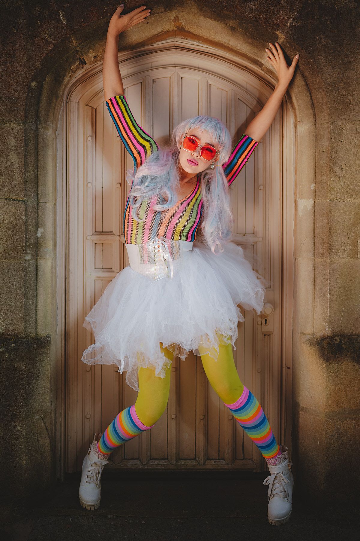 Woman in rainbow outfit with white tutu, yellow tights and roller skates poses at wooden door.