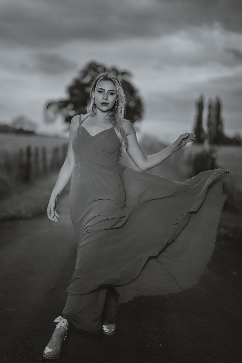 Black and white photo of a blonde woman in a flowing maxi dress walking on a rural road with dramatic cloudy sky.