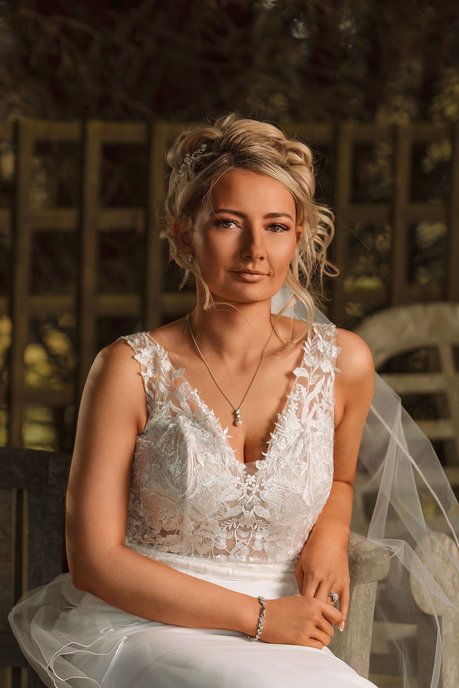 Elegant bride in white lace wedding dress with veil, wearing jewelry, seated outdoors in soft natural light.