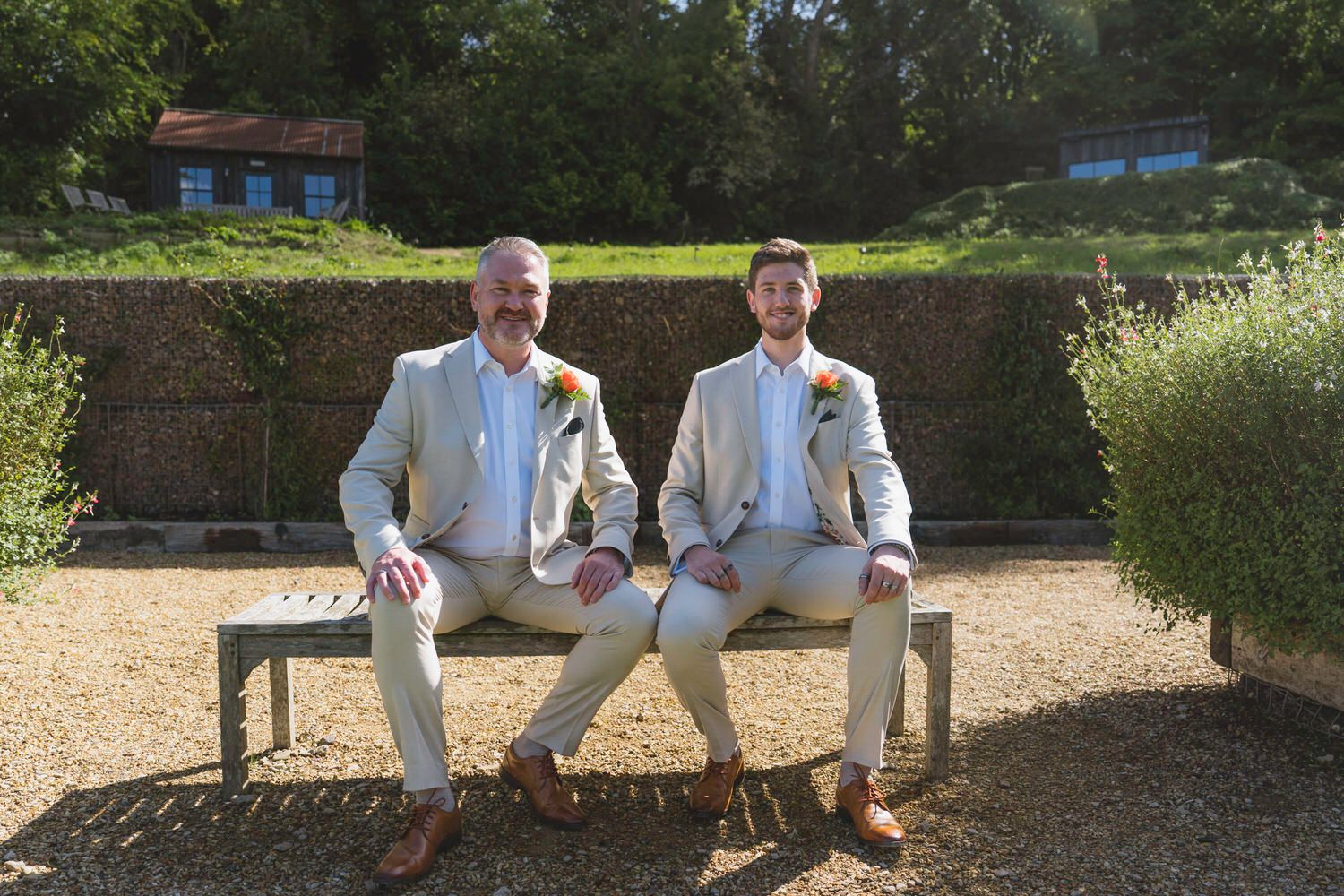 Two grooms in matching beige suits with boutonnieres sit smiling on a bench in a sunny garden setting.