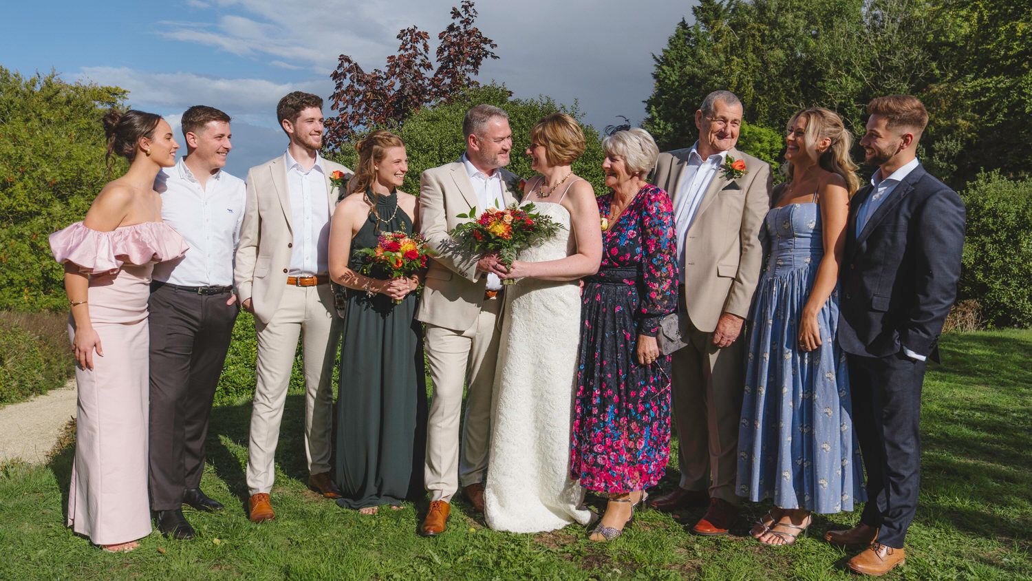 Wedding party poses outdoors on green lawn, bride holding colorful bouquet surrounded by family in elegant attire.