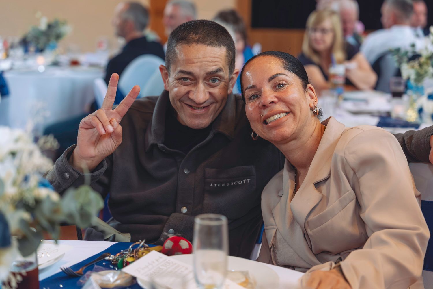 Two smiling people pose at a banquet table, man making peace sign, woman in beige blazer, floral centerpieces in background.