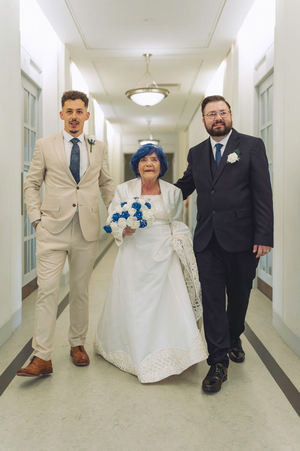 Elderly bride in white dress with blue bouquet escorted by two suited men down hallway.