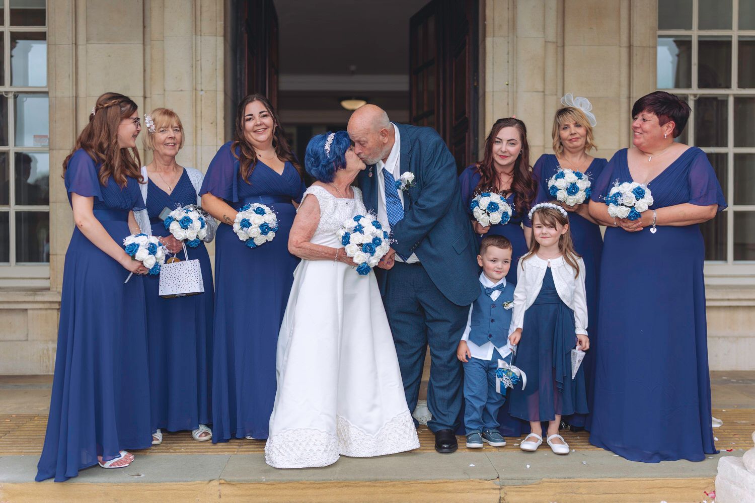 Bride and groom kissing surrounded by bridesmaids in royal blue dresses holding white and blue bouquets at a wedding venue.