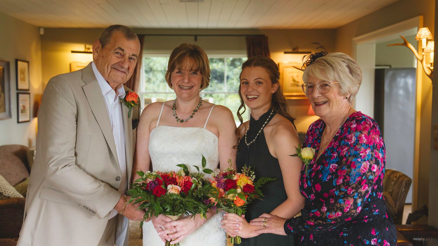 Bride holding colorful bouquet poses with family including parents and bridesmaid indoors at wedding celebration.