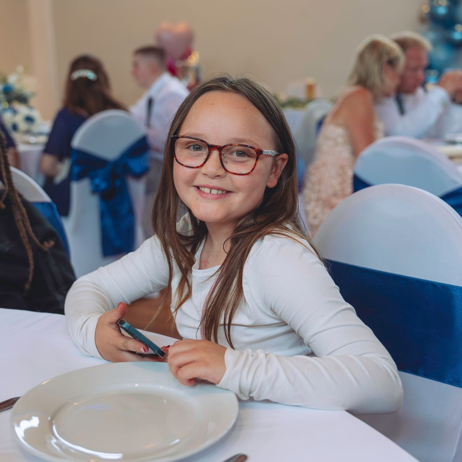 Young girl with glasses smiling at a wedding reception table with a white plate and blue chair covers.