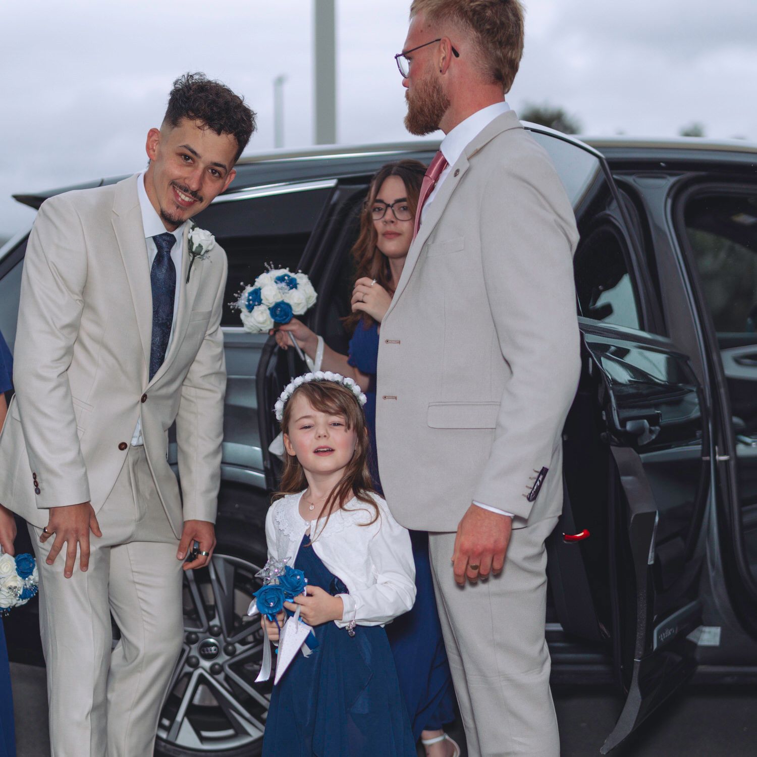 Two groomsmen in beige suits welcome a flower girl in navy blue at a wedding car arrival.