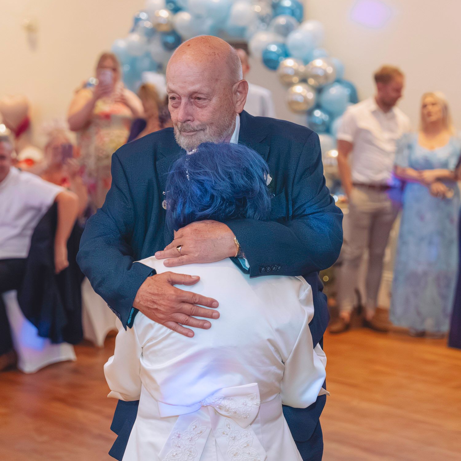 Elderly man in blue suit sharing a heartfelt hug on a dance floor at a wedding reception with balloon decor.