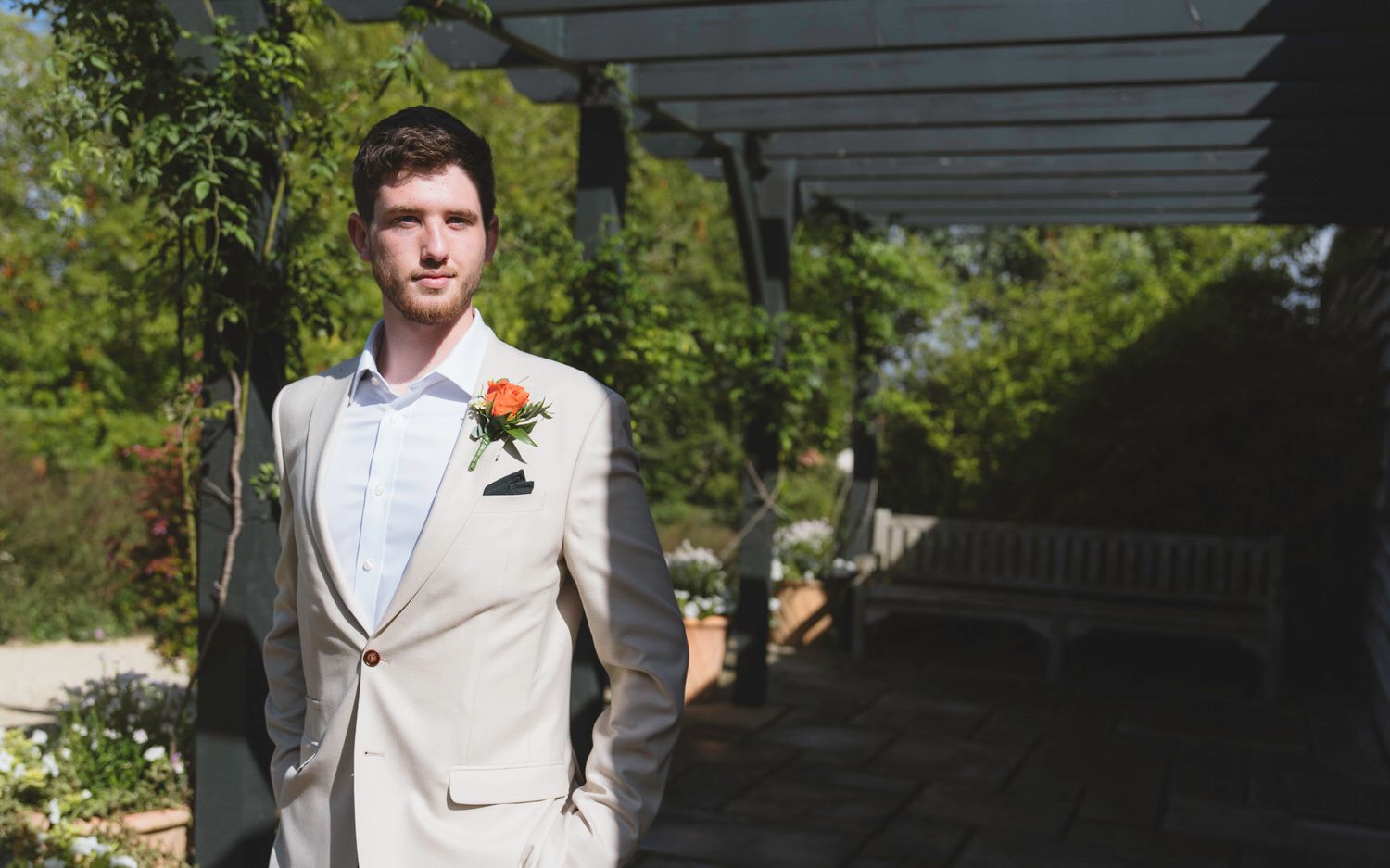 Groom in beige suit with orange boutonniere standing outdoors at garden wedding venue.