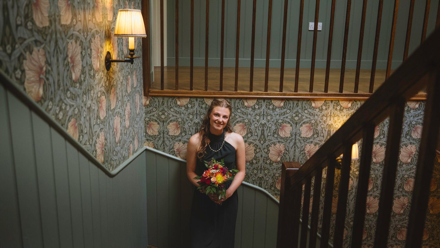 Woman holding colorful bouquet on staircase with elegant floral wallpaper and warm wall lamp lighting.