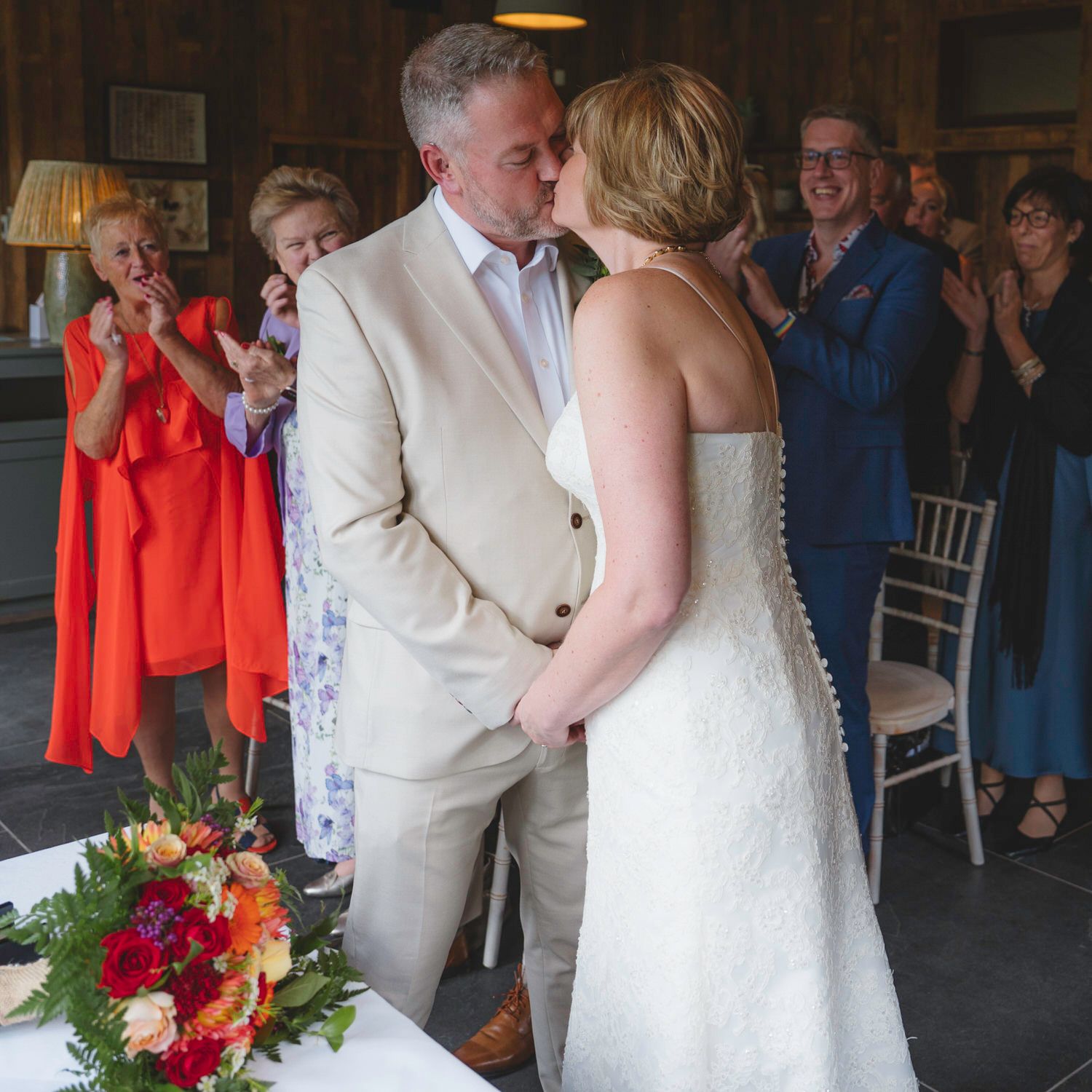Bride and groom share a first kiss at wedding ceremony while guests applaud in background.
