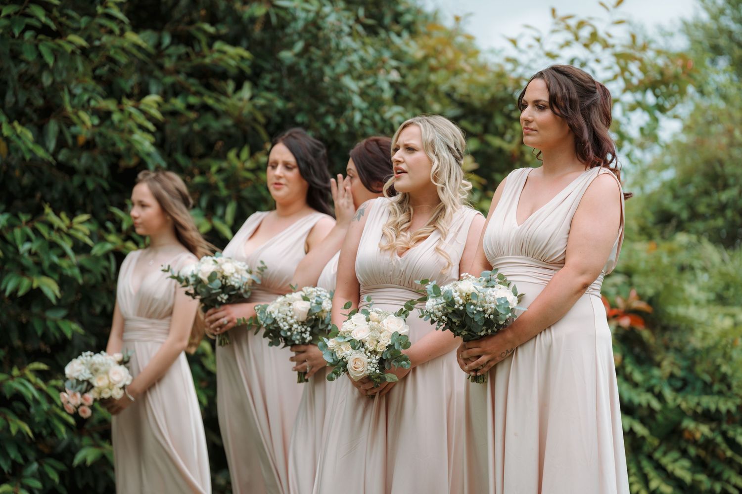 Four bridesmaids in champagne gowns holding white floral bouquets stand outdoors surrounded by lush green foliage.