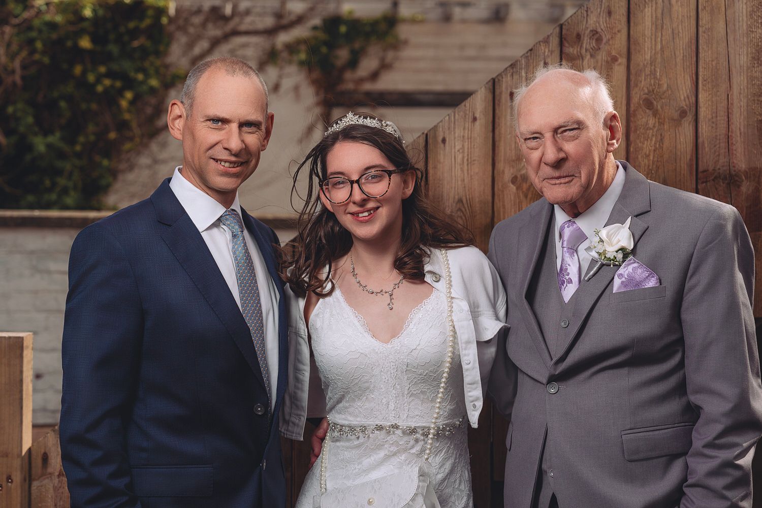 Bride in white dress and tiara poses with two men in suits at a wedding celebration outdoors.
