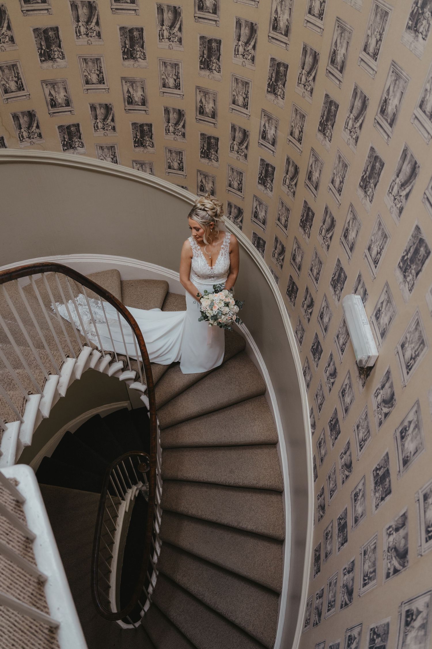 Bride in white gown holding bouquet descends elegant spiral staircase with patterned wallpaper in a grand venue.